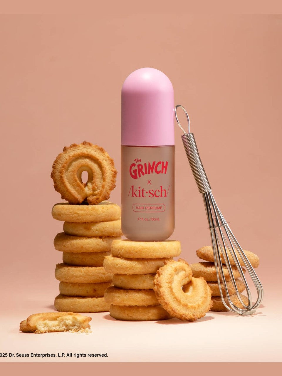 Pink bottle of 'The Grinch' x 'kitsch' hair perfume with cookies and a whisk on a beige background