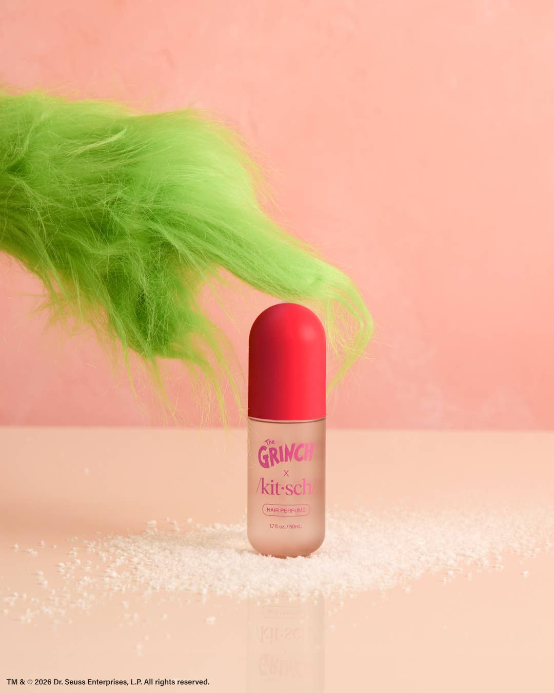 A bottle of Dr. G's Grinch & Kitchez lip gloss stands on a pink surface scattered with white glitter, with a bright neon green fluffy tail-like shape against a soft pink background.