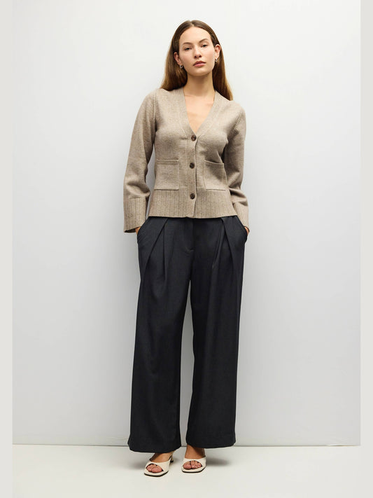 A woman stands against a light gray studio wall wearing a beige knit cardigan with front buttons and dark wide-leg trousers, paired with white slide sandals.